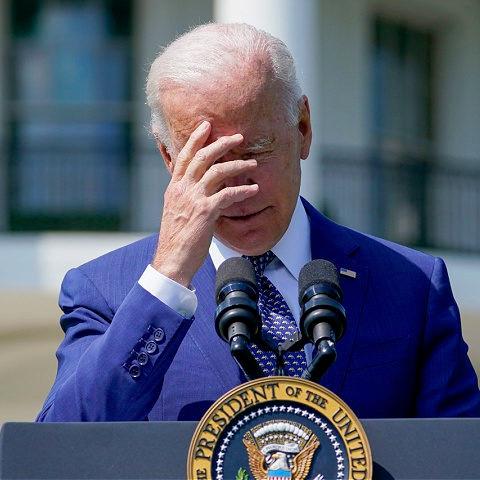 A photo of Joe Biden while standing at a lecturn with a Presidential Seal with a facepalm expression.